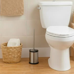 Brushed stainless steel toilet brush with black trim and chrome handle beside white toilet in bathroom with woven basket on wood floor