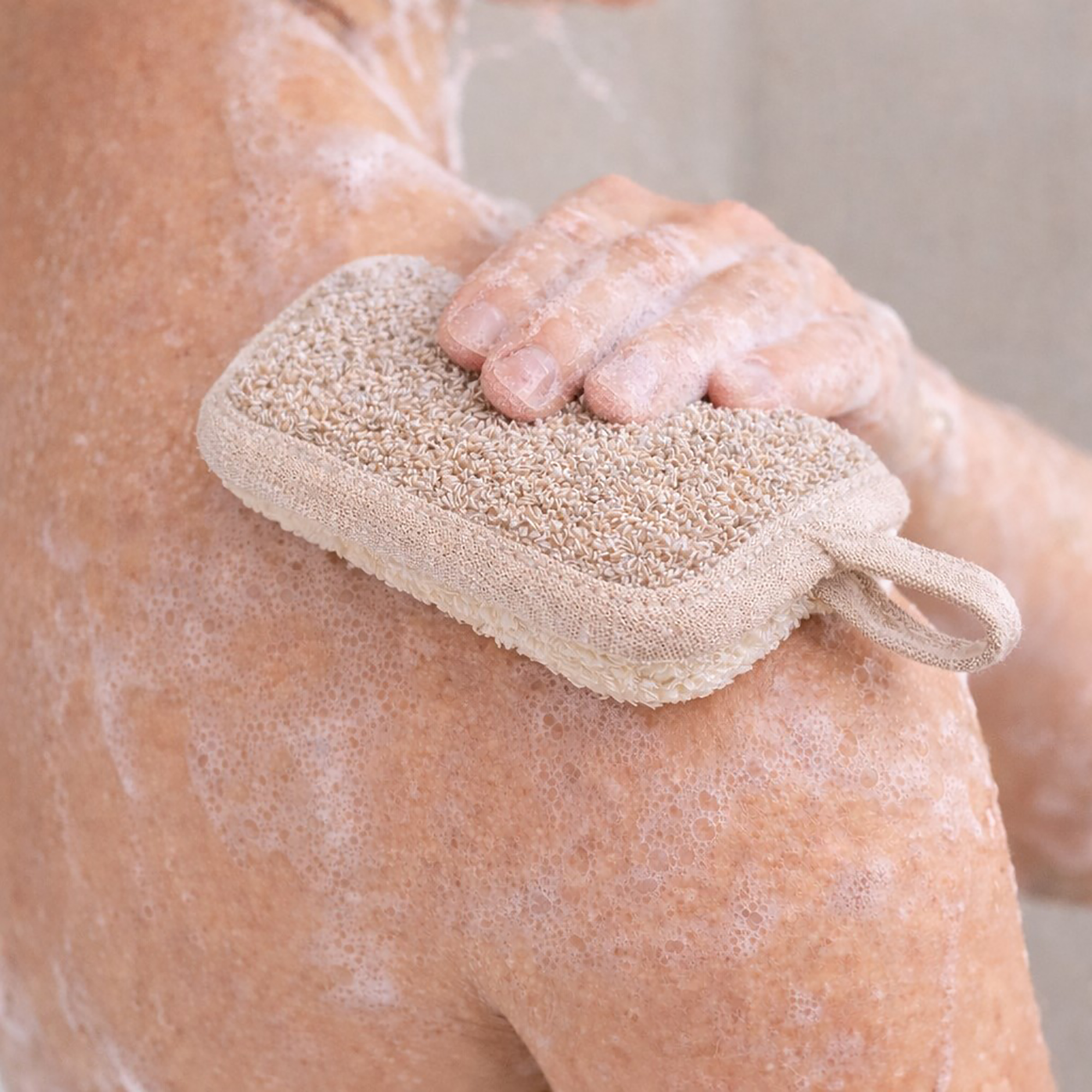 Person using beige dual-texture exfoliating pad with fabric loop on soapy shoulder