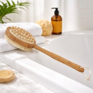 Dual-sided wooden body brush with natural bristles and massage nodules on a long bamboo handle, resting on a white bathtub ledge with a towel, sponge, and amber bottle nearby.