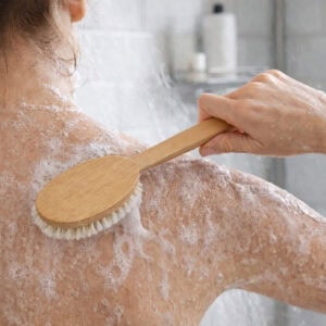 Woman using a long-handled wooden bath brush to scrub her soapy back in the shower.