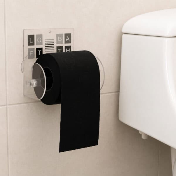 Loft Bath clear acrylic toilet paper holder suction-mounted on beige tile beside a toilet tank, holding a black roll with paper hanging down. Graphic insert spells "LOFT BATH" in black, grey, and white lettered tiles.
