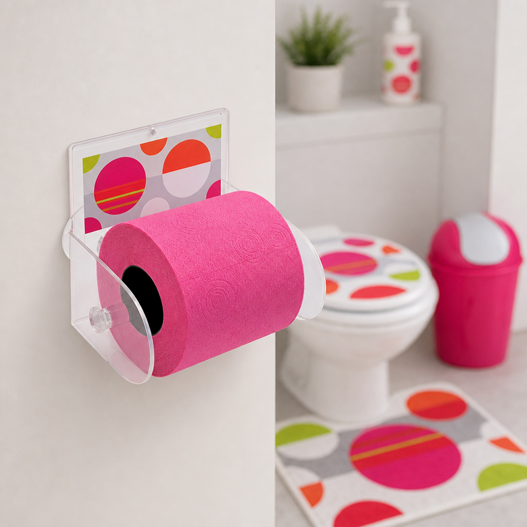 Eclats clear acrylic toilet paper holder mounted on wall holding a hot pink roll, with toilet, matching polka-dot seat cover, pink swing-lid bin, and coordinating bath mat in the background.