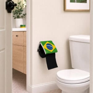 Brazil flag print toilet paper holder mounted on a wall in a styled bathroom, holding a black roll under a flip-up Brazilian flag cover, with a vanity, eucalyptus, and framed art nearby.