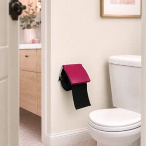 Fuchsia cover toilet paper holder mounted on a wall in a styled bathroom, holding a black roll under a flip-up fuchsia cover, with a vanity, eucalyptus, and framed art nearby.