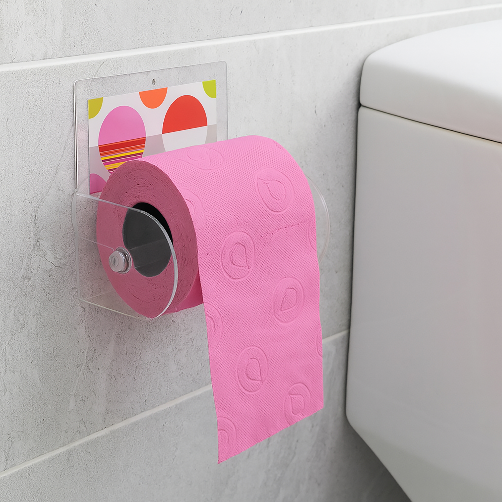 Eclats clear acrylic toilet paper holder suction-mounted on grey tile beside a toilet tank, holding a pink roll with paper hanging down.