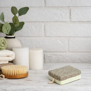 Beige and white dual-texture exfoliating pad with spa accessories including candles, brush, and towels