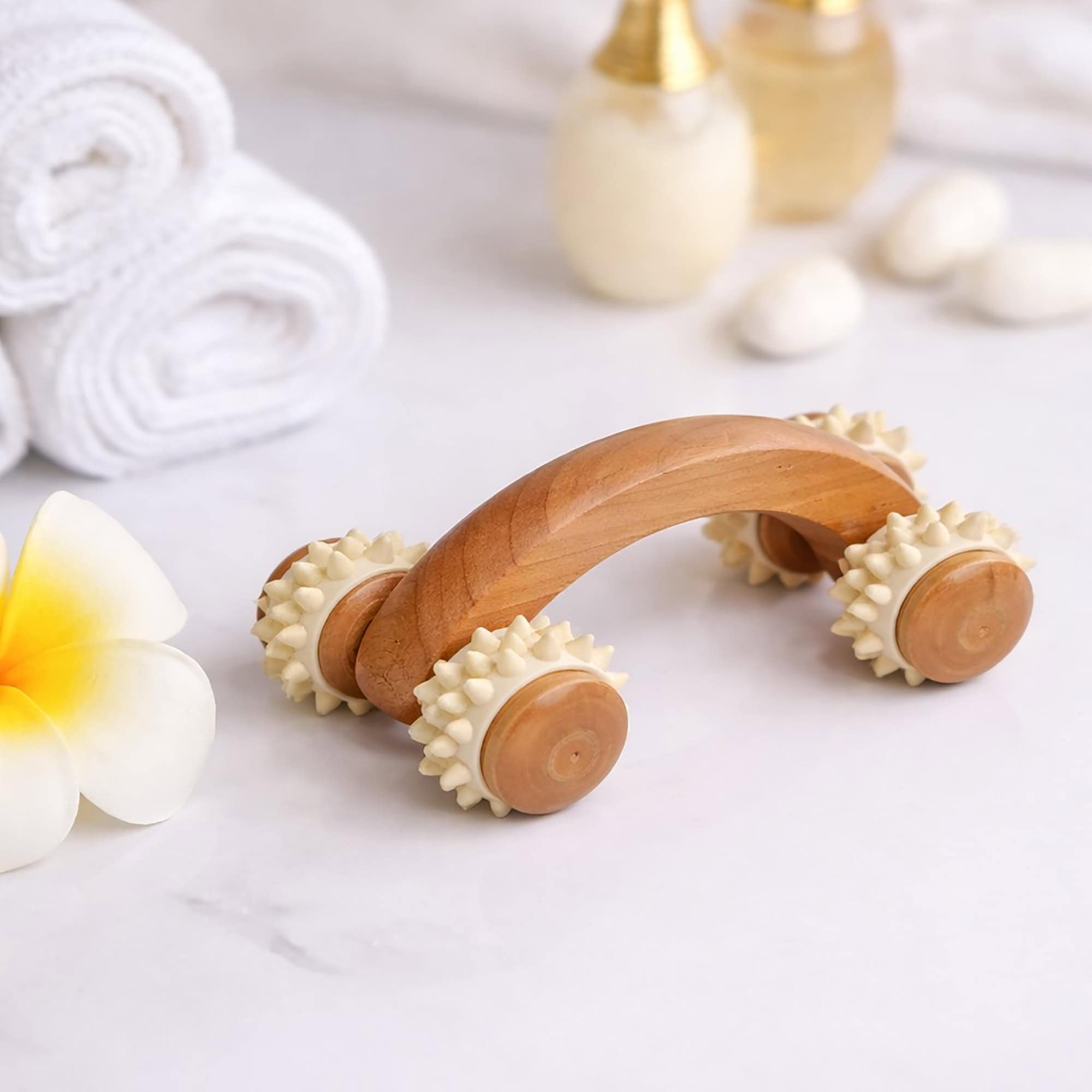 Wooden massage roller styled on white fabric with spa accessories including white flowers and golden wellness items