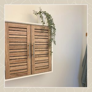 "Close-up of oak louvered wall cabinet with stainless steel handles, topped with a trailing eucalyptus plant"