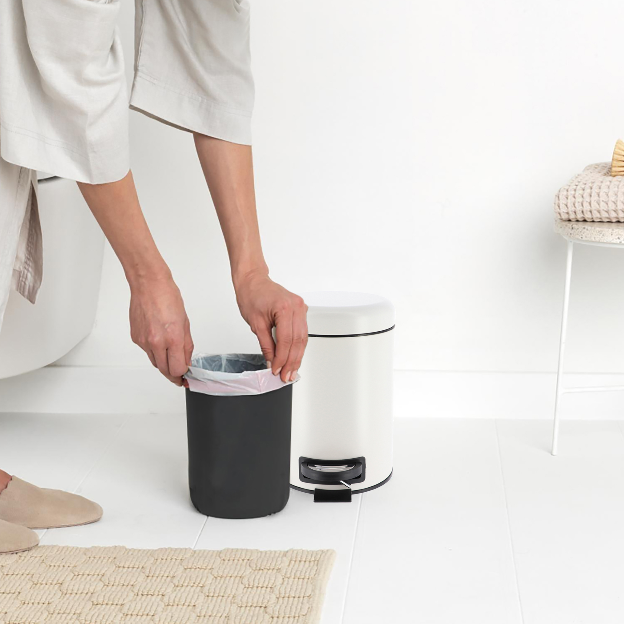 Person in white robe placing pink trash bag liner into small white step can
