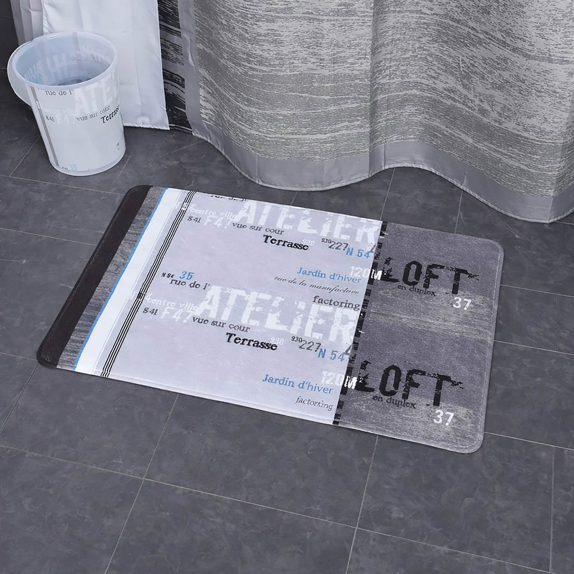 Close-up of Atelier Loft bath mat with French text design on dark tile floor beside shower curtain