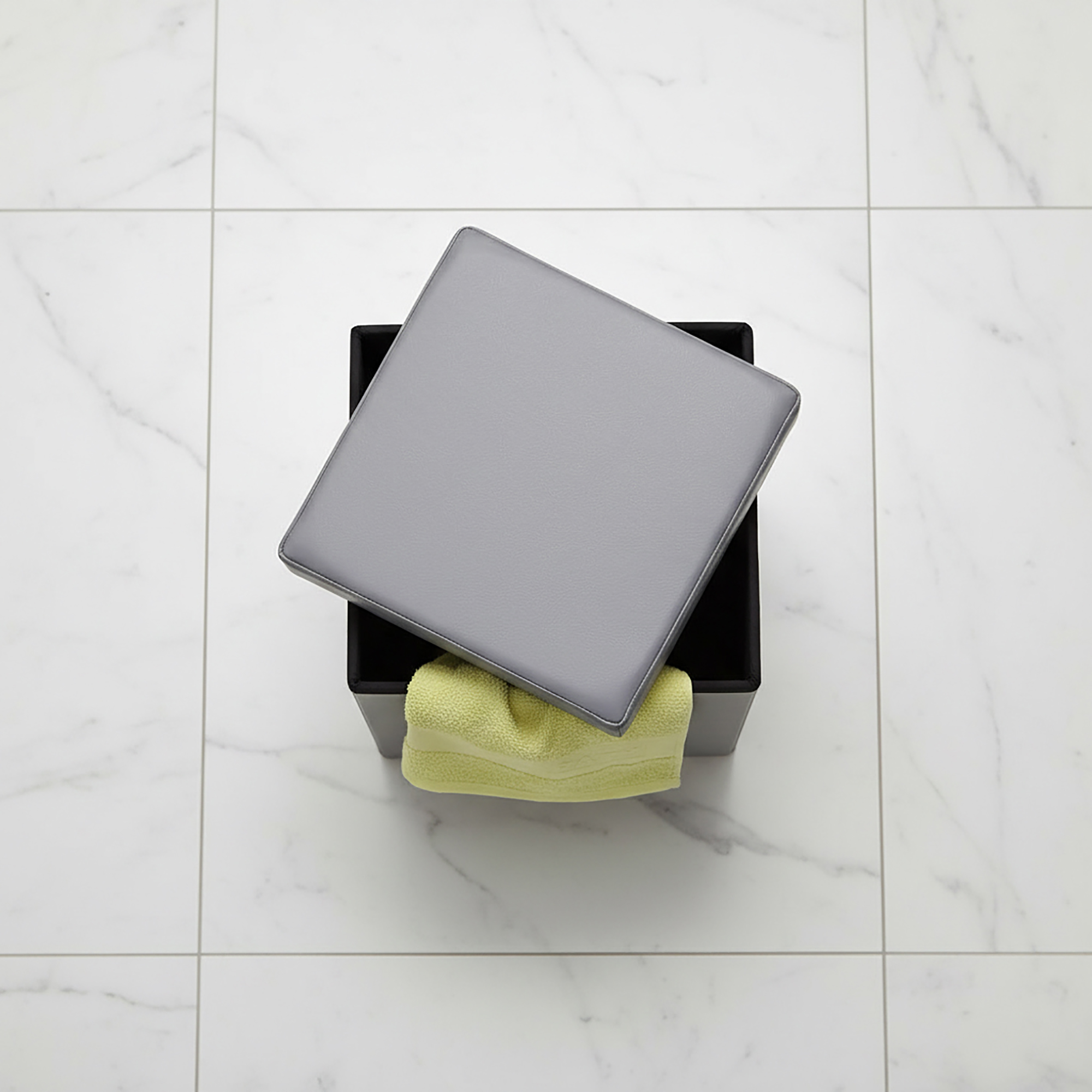 "Overhead view of gray storage ottoman with lid ajar revealing yellow towel inside, on marble floor"
