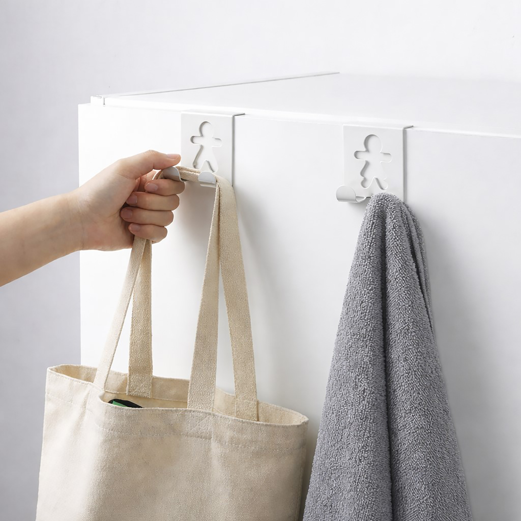 "Hand removing tote bag from white figure-shaped cabinet hook beside second hook holding gray towel"