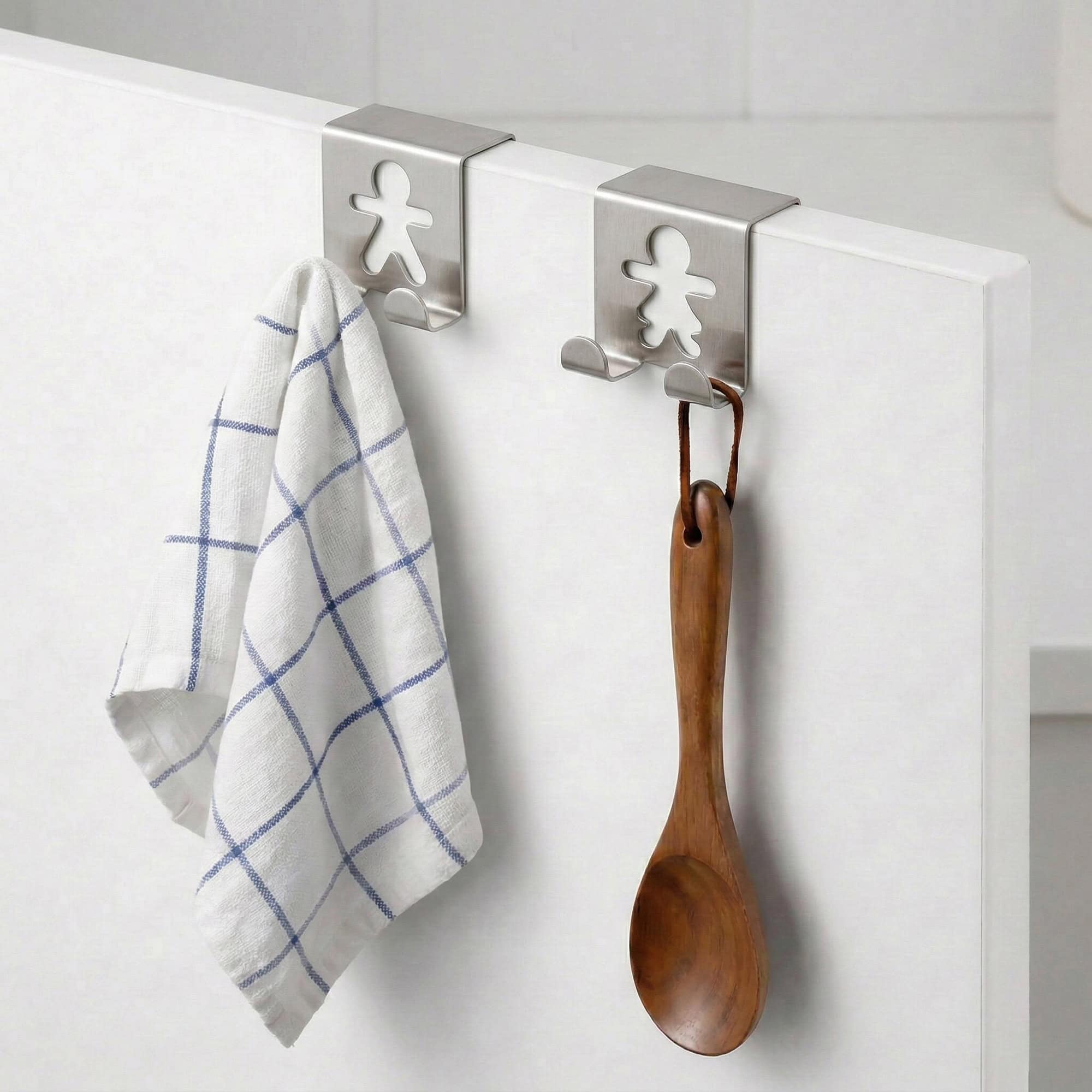 "Two stainless steel figure hooks on white cabinet holding blue plaid dish towel and wooden spoon"