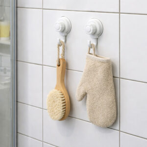 Two suction cup hooks on shower tiles holding a wooden body brush and exfoliating mitt