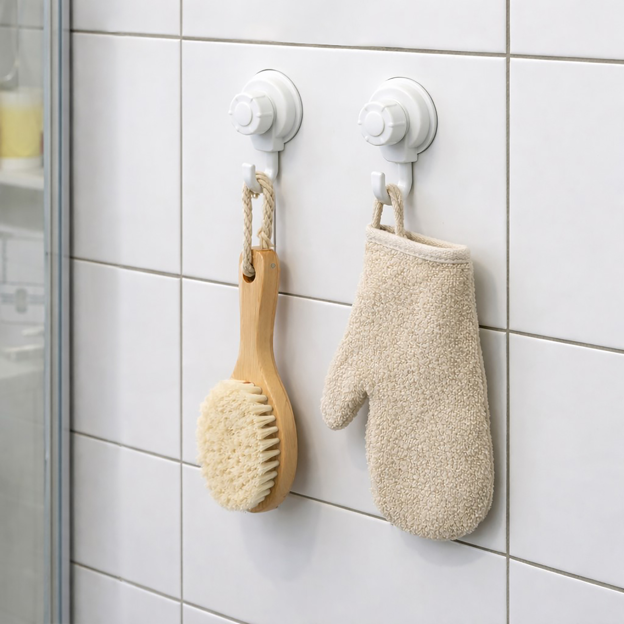 Two suction cup hooks on shower tiles holding a wooden body brush and exfoliating mitt