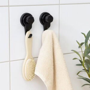 Two black suction cup hooks on white tiles holding a hairbrush and cream towel