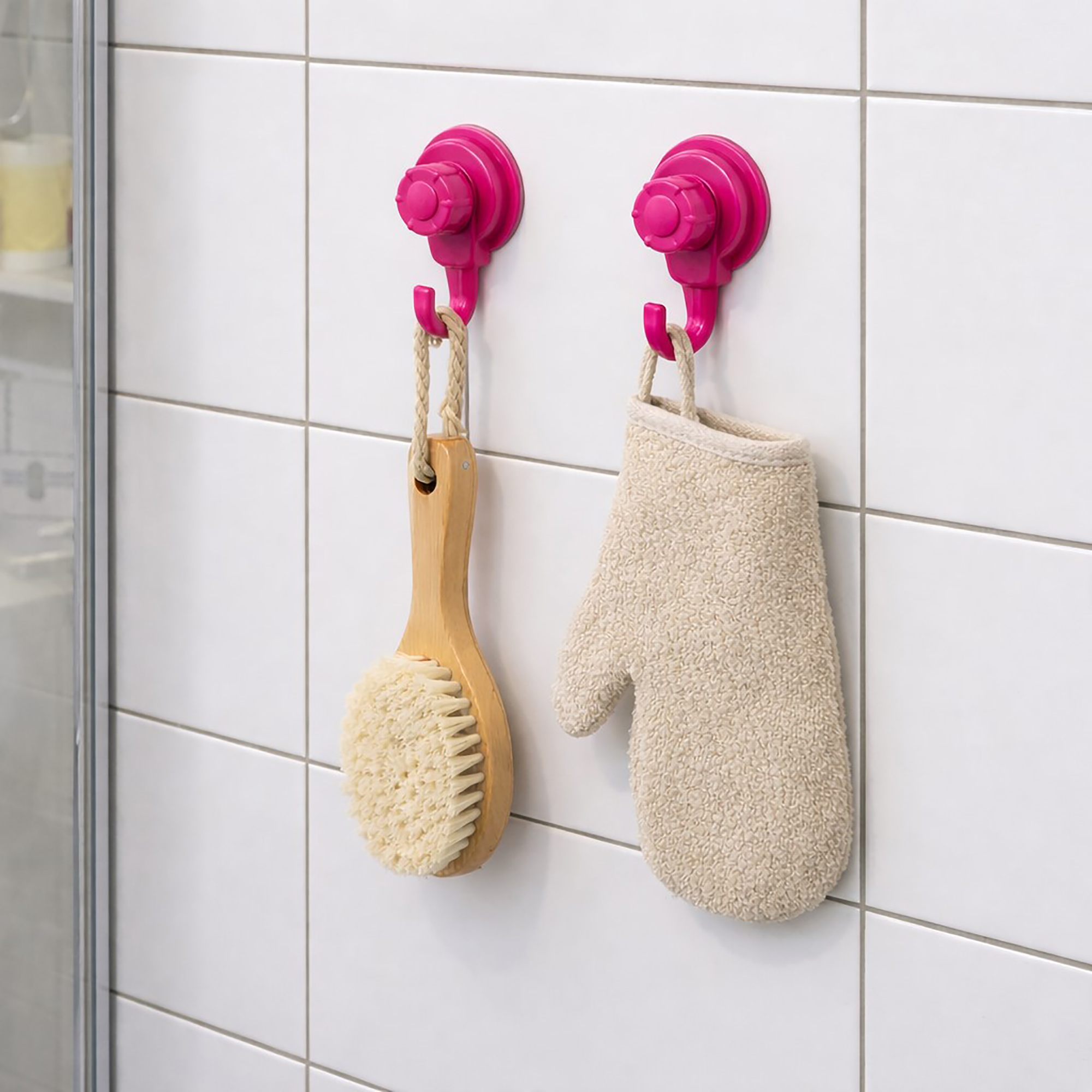 Two hot pink suction cup hooks on shower tiles holding a wooden body brush and exfoliating mitt