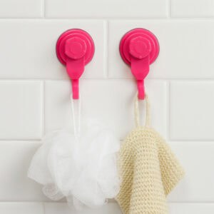 Two hot pink suction cup hooks on white subway tiles holding a white bath puff and natural exfoliating mitt