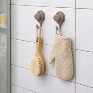Two tan suction cup hooks on shower tiles holding a wooden body brush and exfoliating mitt