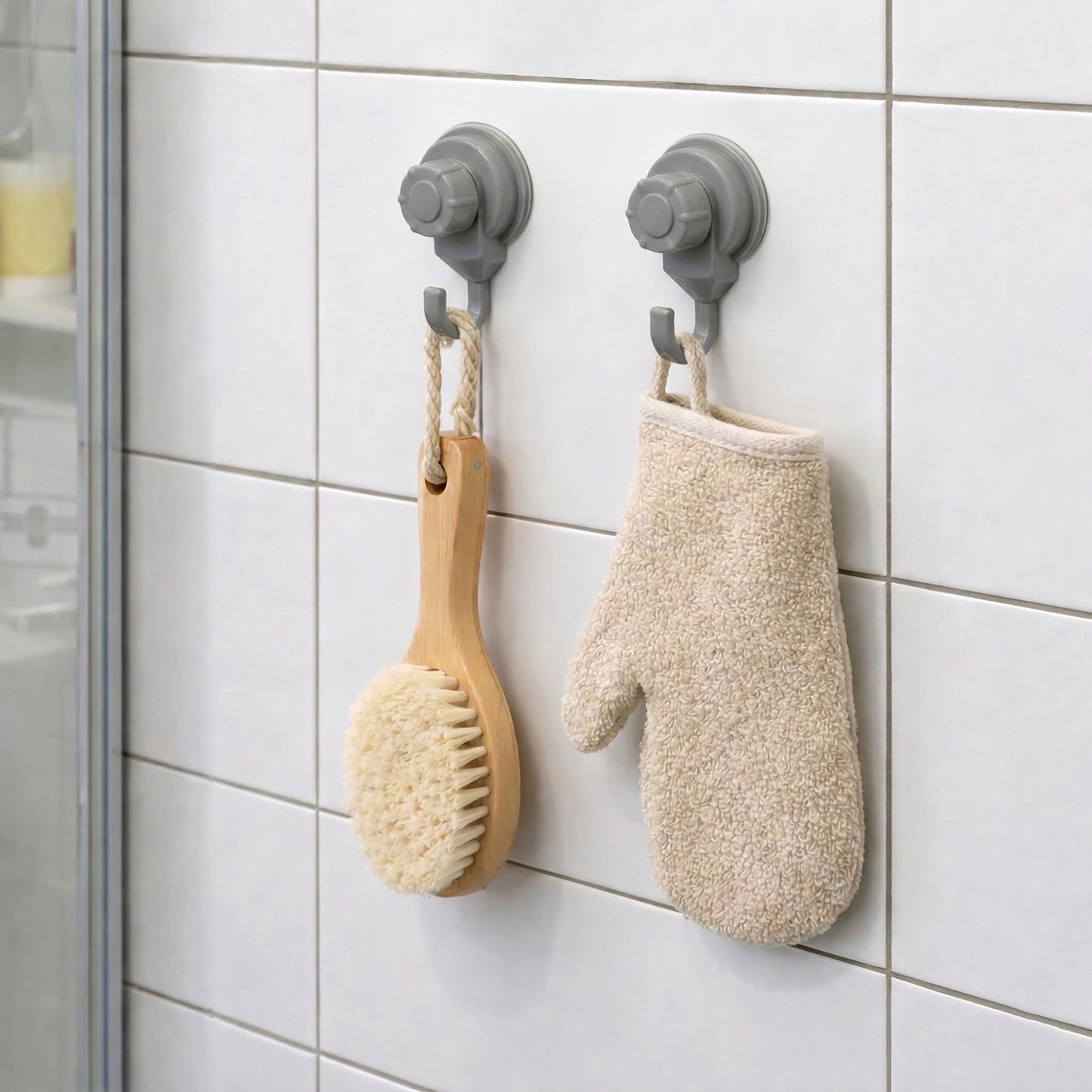 Two gray suction cup hooks on shower tiles holding a wooden body brush and exfoliating mitt