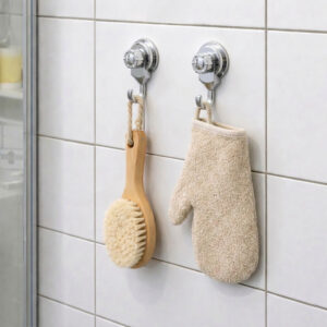 Two chrome suction cup hooks on shower tiles holding a wooden body brush and exfoliating mitt