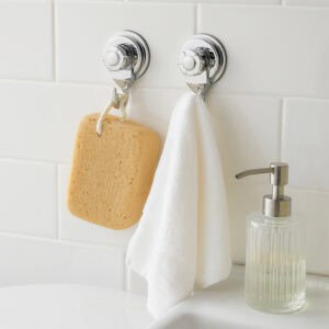Two chrome suction cup hooks on white subway tiles holding a natural sponge and white towel, with glass soap dispenser nearby