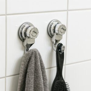 Two chrome suction cup hooks on white tiles holding a gray towel and black hairbrush