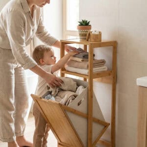 Adult and toddler placing laundry and folded towels into open bamboo hamper cabinet in a bright bathroom