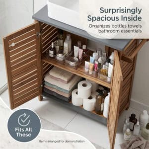 Acacia wood under-sink cabinet open with doors spread wide, interior shelves holding skincare bottles, folded towels, toilet paper rolls, and cleaning supplies