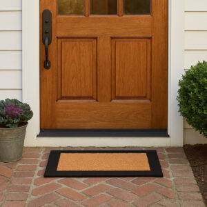 Natural coir door mat with black border on a brick porch in front of a wooden front door