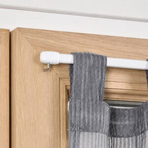 Close up of a white cafe rod resting on a chrome screw in L hook mounted to a wood window frame, holding a sheer gray tier curtain.