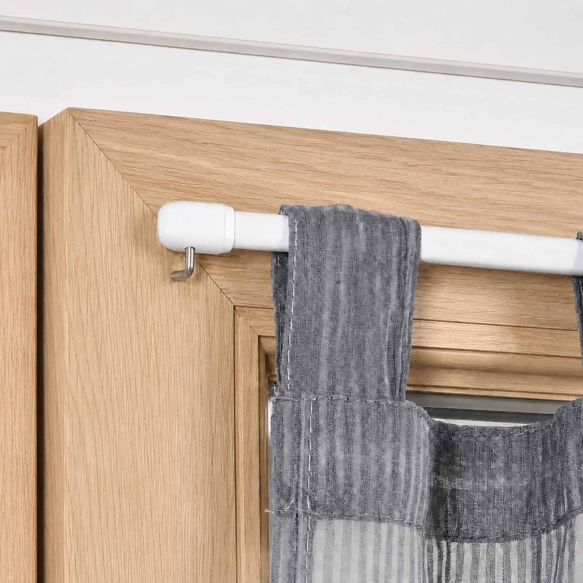 Close up of a white cafe rod resting on a chrome screw in L hook mounted to a wood window frame, holding a sheer gray tier curtain.
