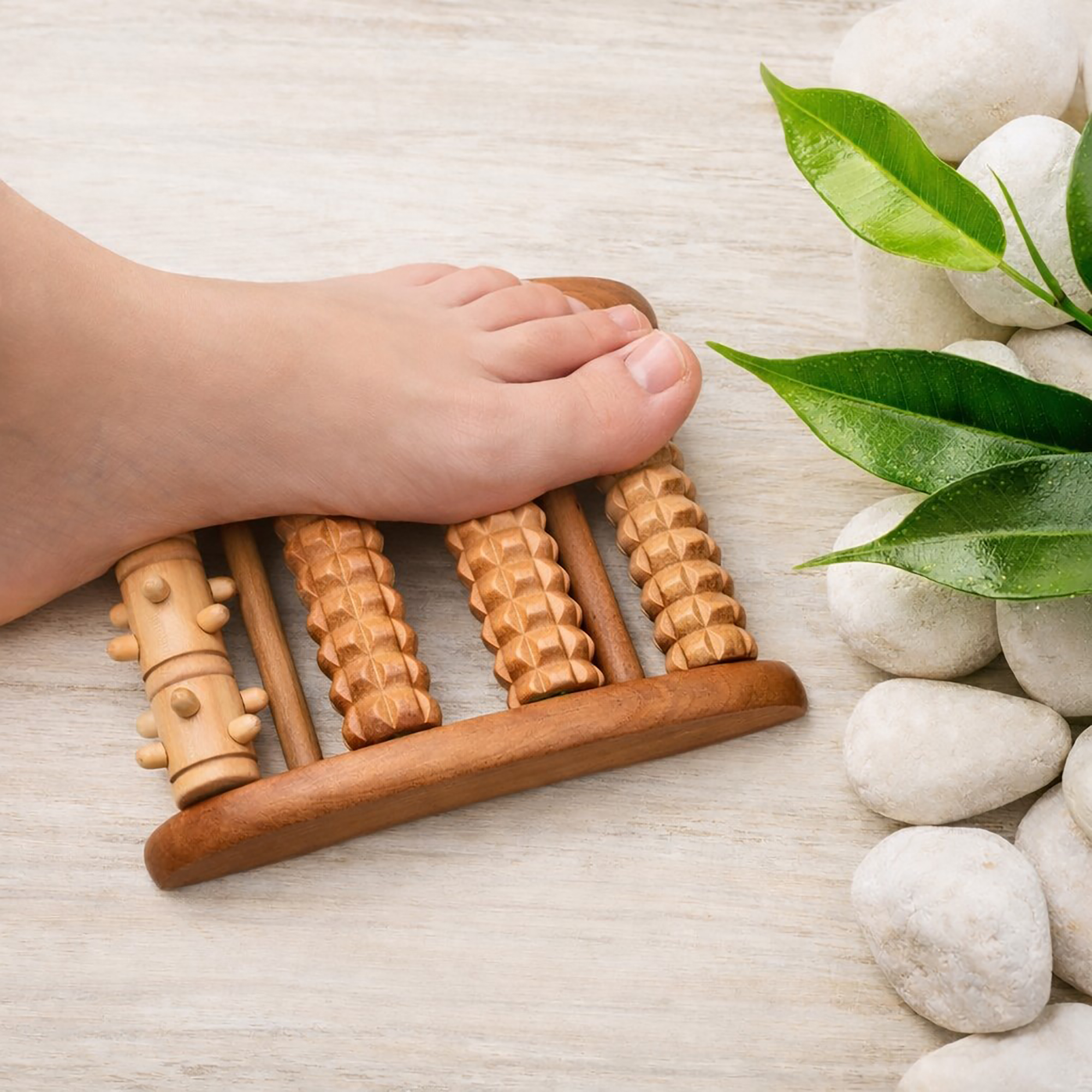 Bare foot pressing down on a wooden foot roller massager with five textured rollers, with white stones and green leaves alongside.