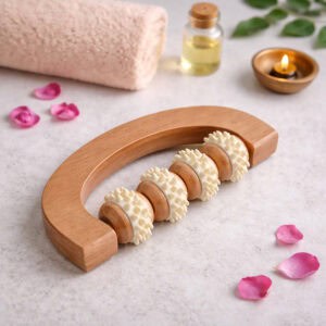 Wooden U-shaped massage roller with four textured spiked wheels on a marble surface, surrounded by rose petals, a rolled towel, essential oil, and a tea light candle.