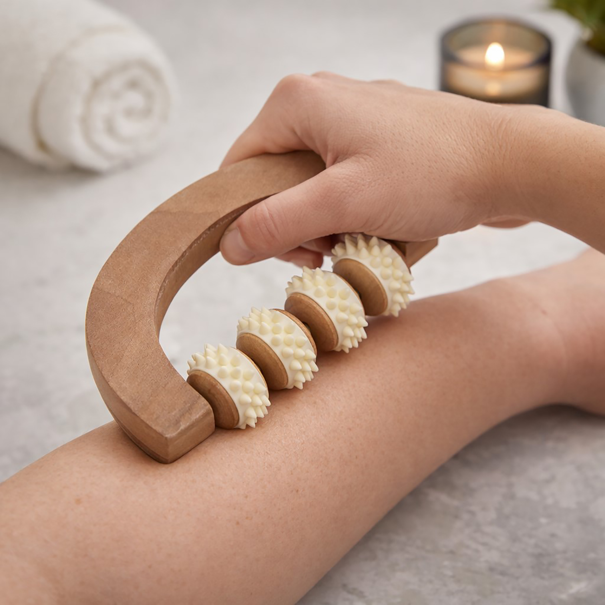 Hand using a wooden U-shaped massage roller with four textured spiked wheels on a person's forearm, with a rolled towel and candle in the background.