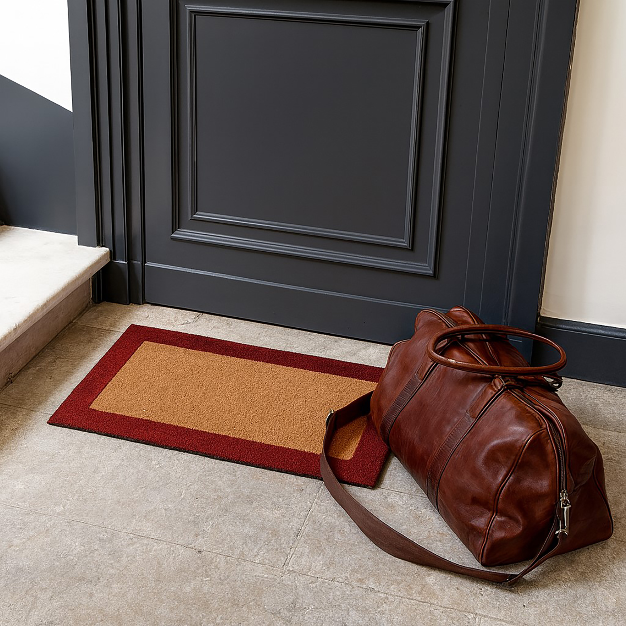 Small red-border coir doormat 24x13 by dark entry door with travel bag