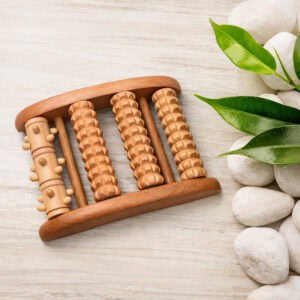 Wooden foot roller massager with five textured rollers on a light wood surface, surrounded by white stones and green leaves.