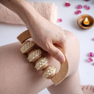 Person using wooden hand massage roller on arm in spa setting with pink flowers, candles, and bath accessories