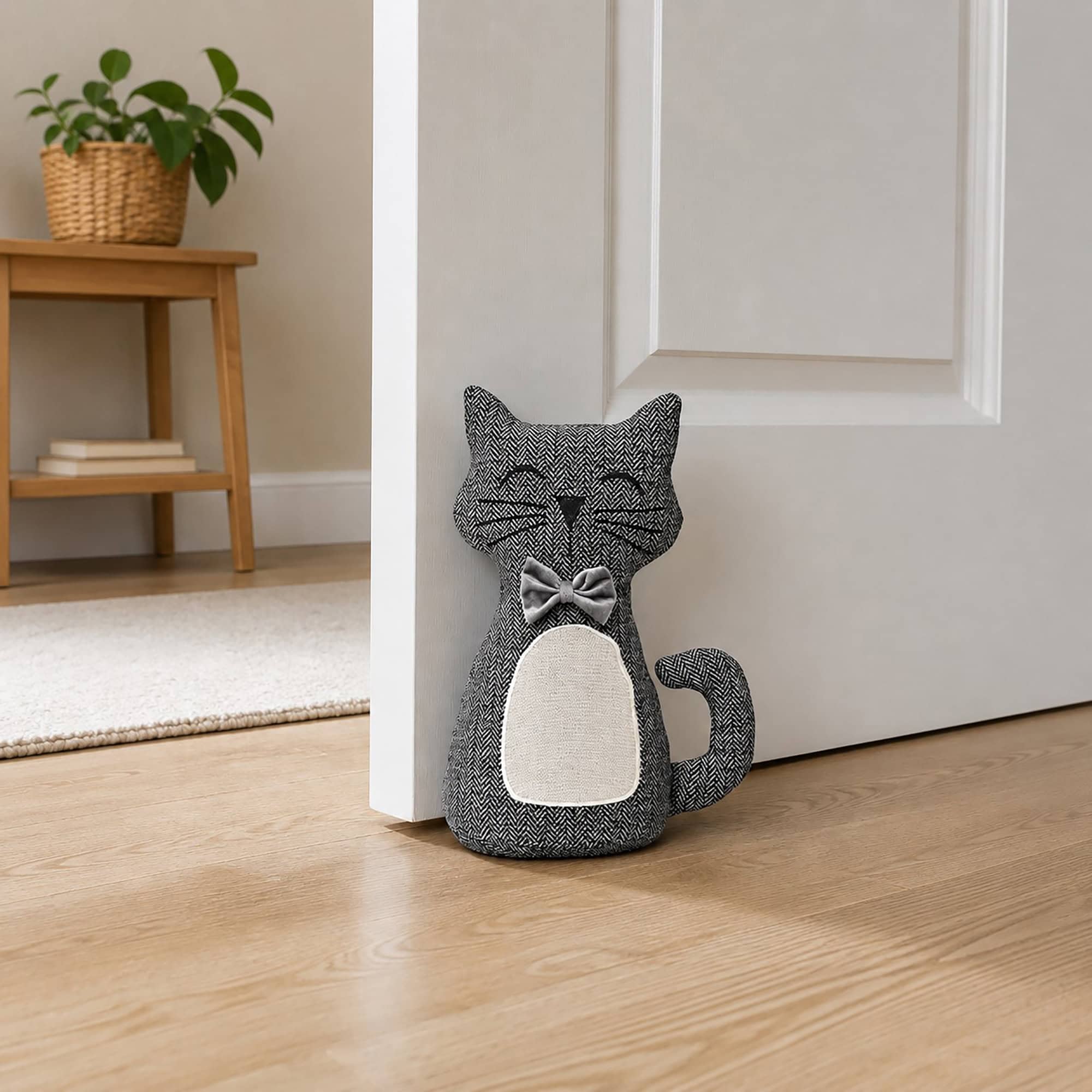 Charcoal cat door stopper propping open a white door, hardwood floor, rug, wooden bench with a potted plant in the background.