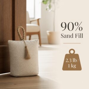 Natural fabric bag door stopper, cream woven cotton with a twisted rope handle and jute tassel, propping open a wood door on hardwood floor, armchair in the background. On-image text: "90% Sand Fill," "2.3 lb / 1 kg."