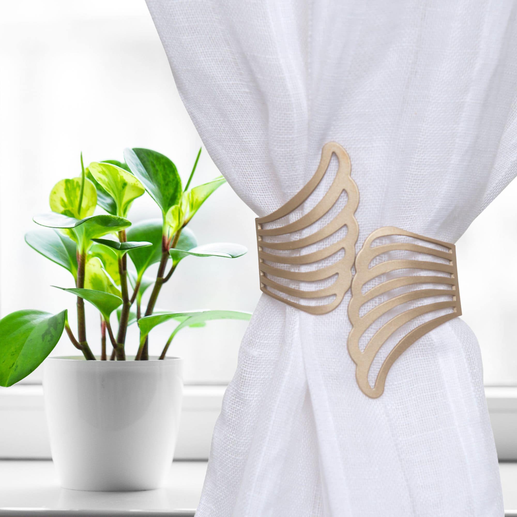 "Gold angel wings tieback on white curtain beside a potted plant on a windowsill"