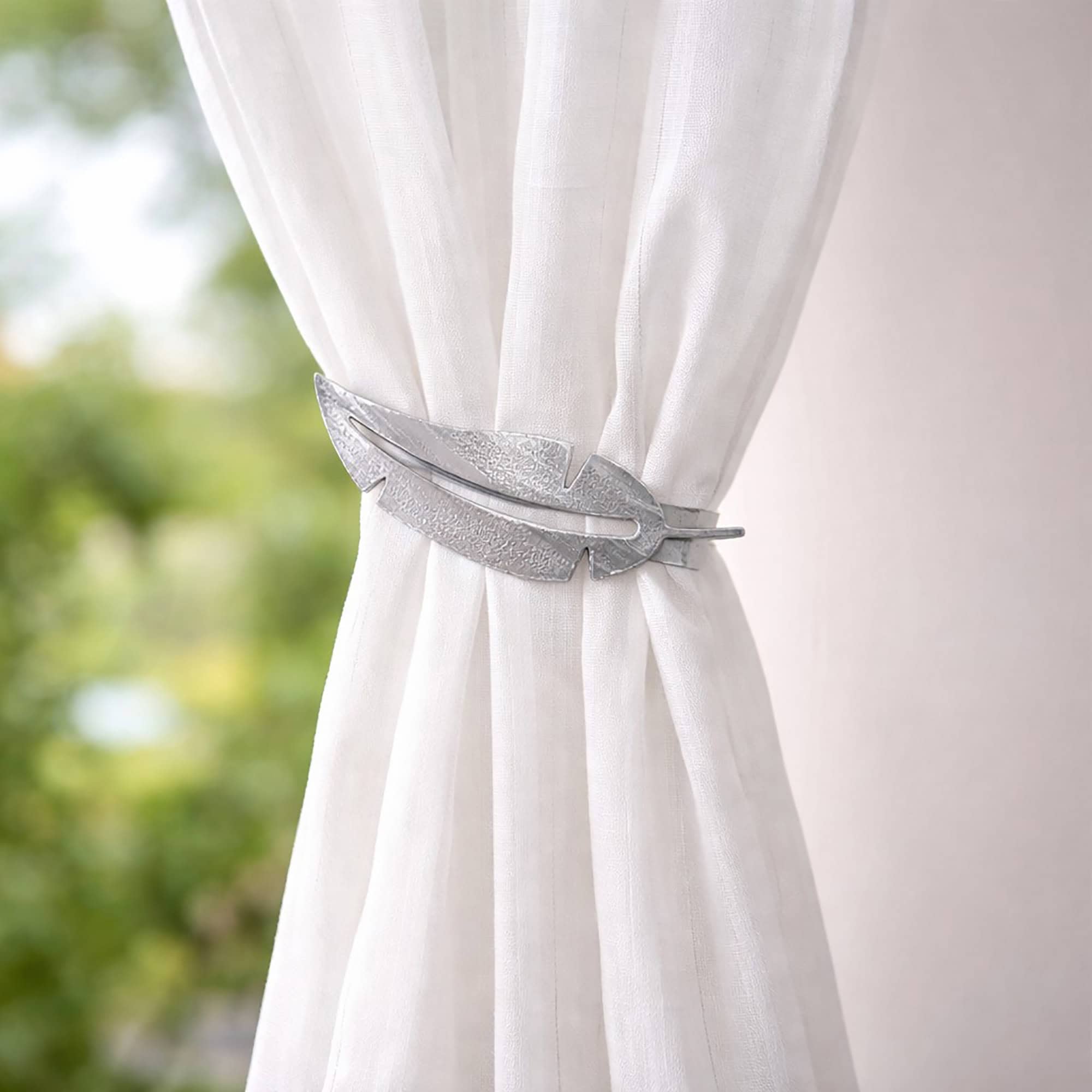 "Grey metal feather tieback on sheer white curtain with green garden backdrop"