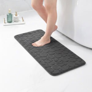 Barefoot stepping on a long dark gray runner mat with pebble texture placed on a bathroom tile floor next to a tub