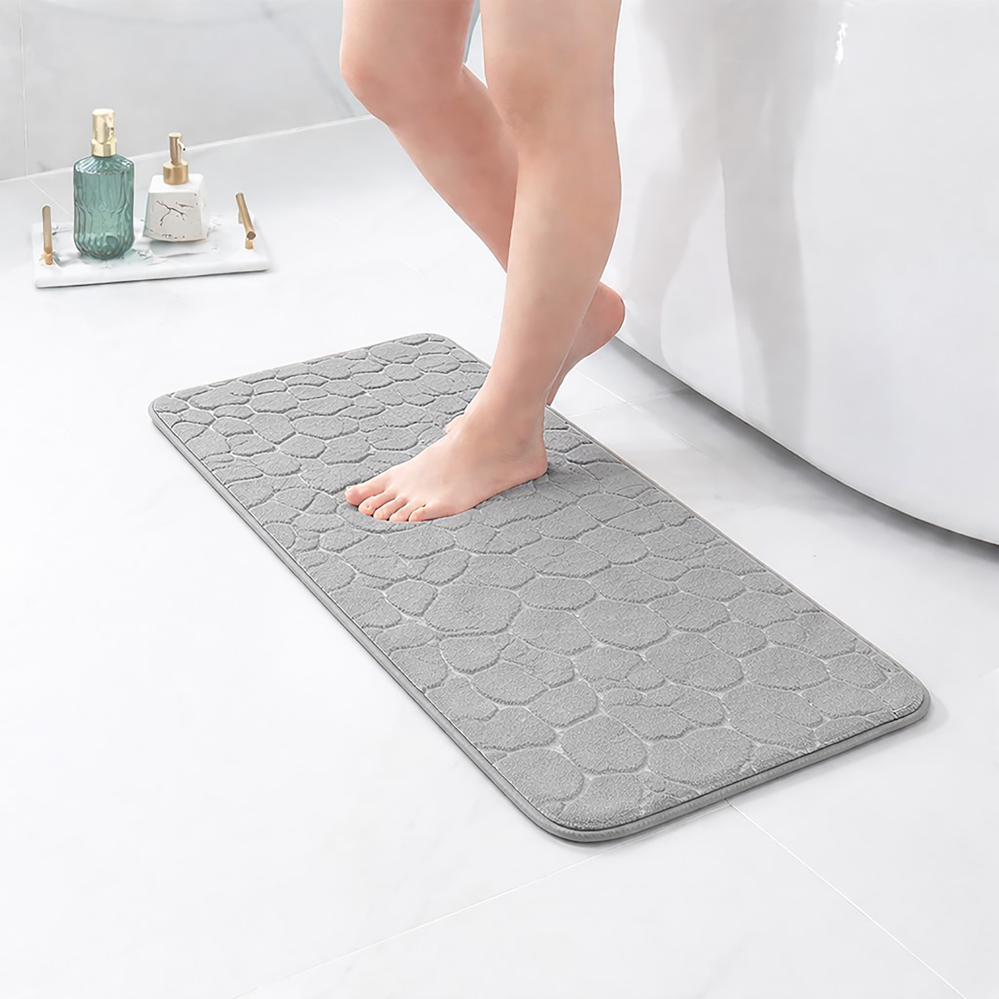 Barefoot stepping on a long light gray runner mat with pebble texture placed on a bathroom tile floor next to a tub