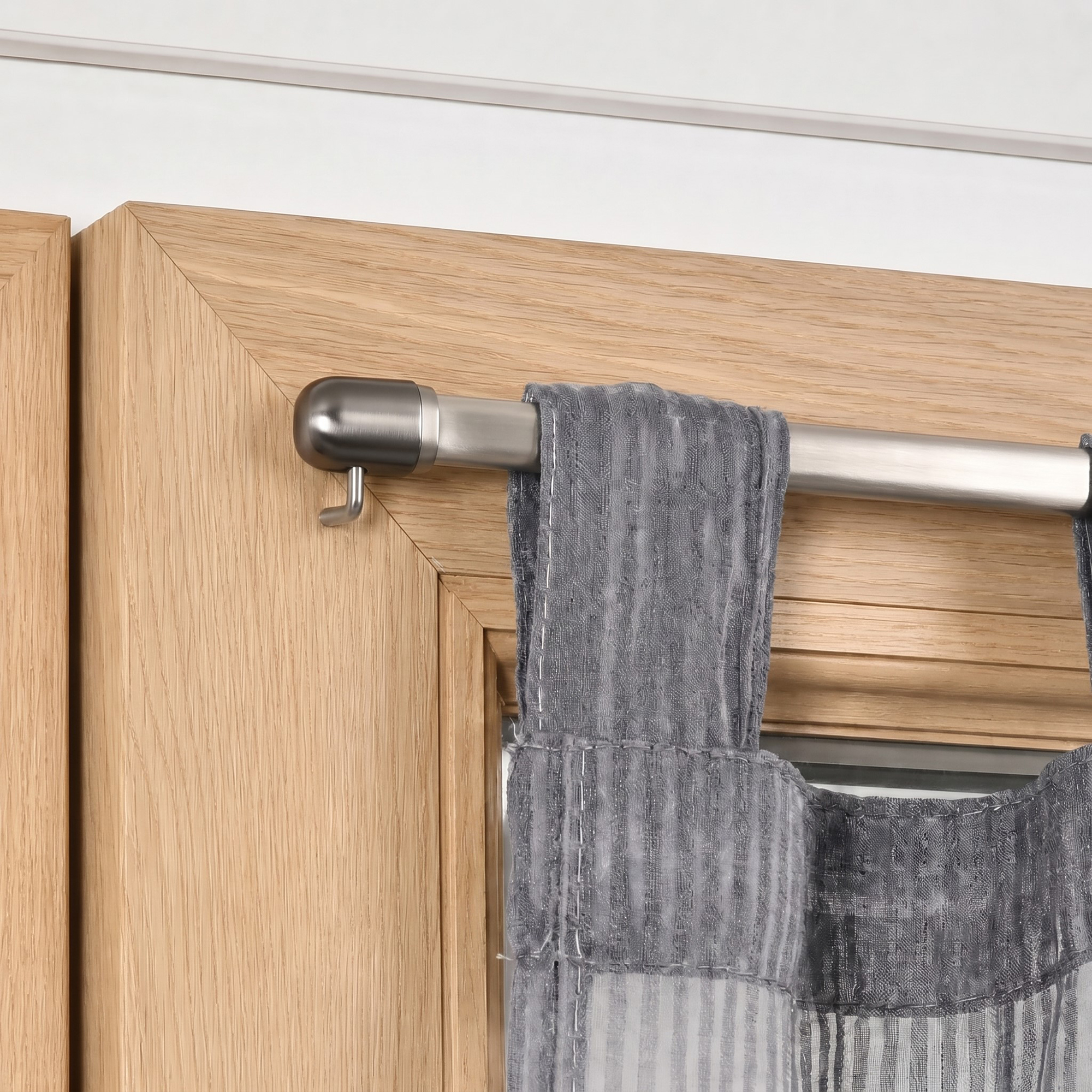 Close-up of a brushed nickel cafe curtain rod resting on a mounting hook attached to a wooden window frame.