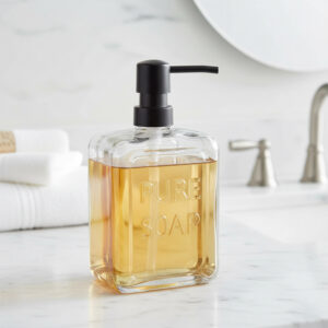 Clear glass soap dispenser Pure Soap 18 fl oz filled with amber soap on a bathroom vanity beside folded white towels