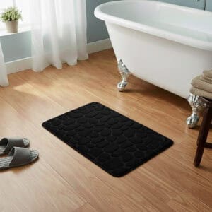 Black memory foam bath mat with embossed pebble stone texture on wooden floor beside clawfoot tub
