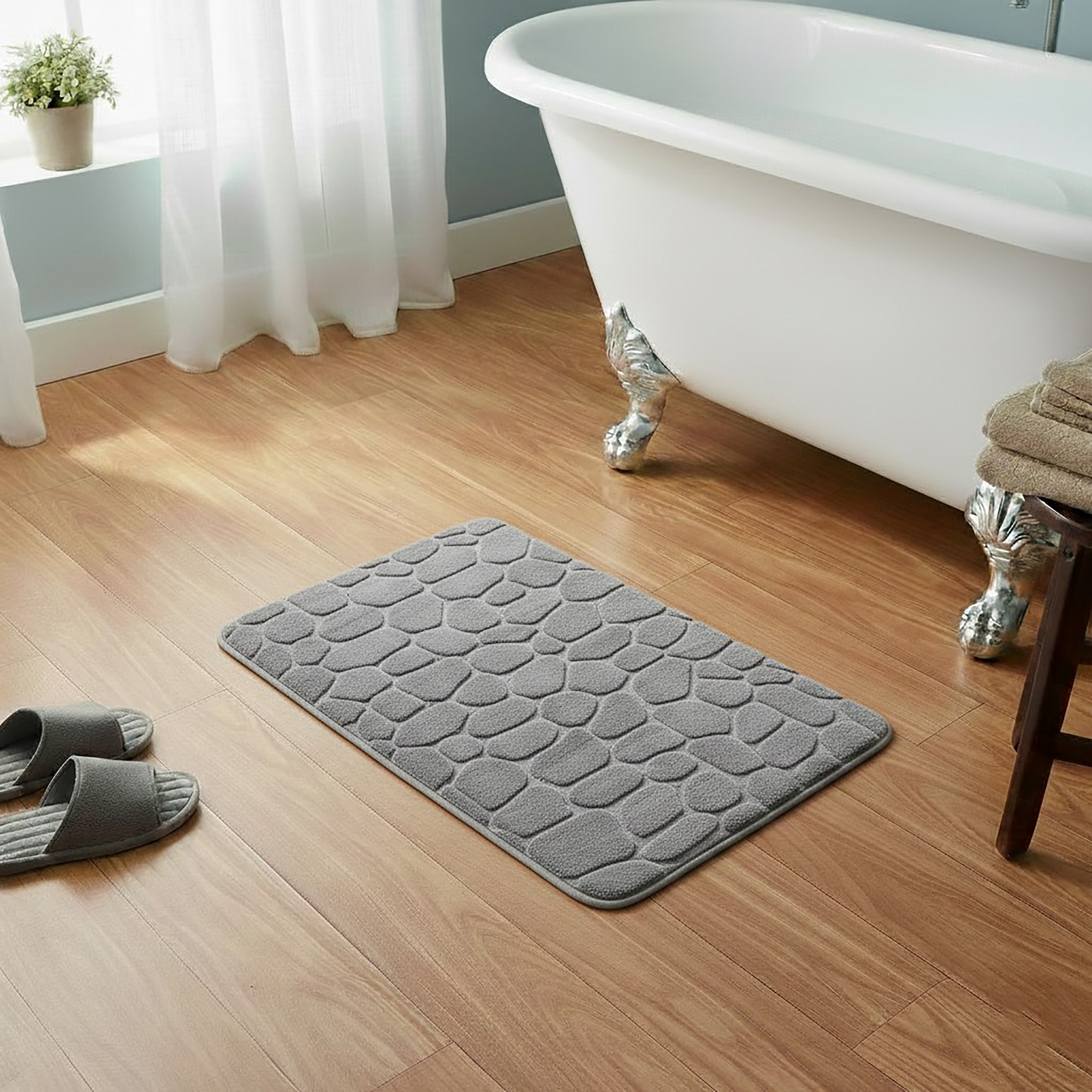 Light gray memory foam bath mat with embossed pebble stone texture on wooden floor beside clawfoot tub