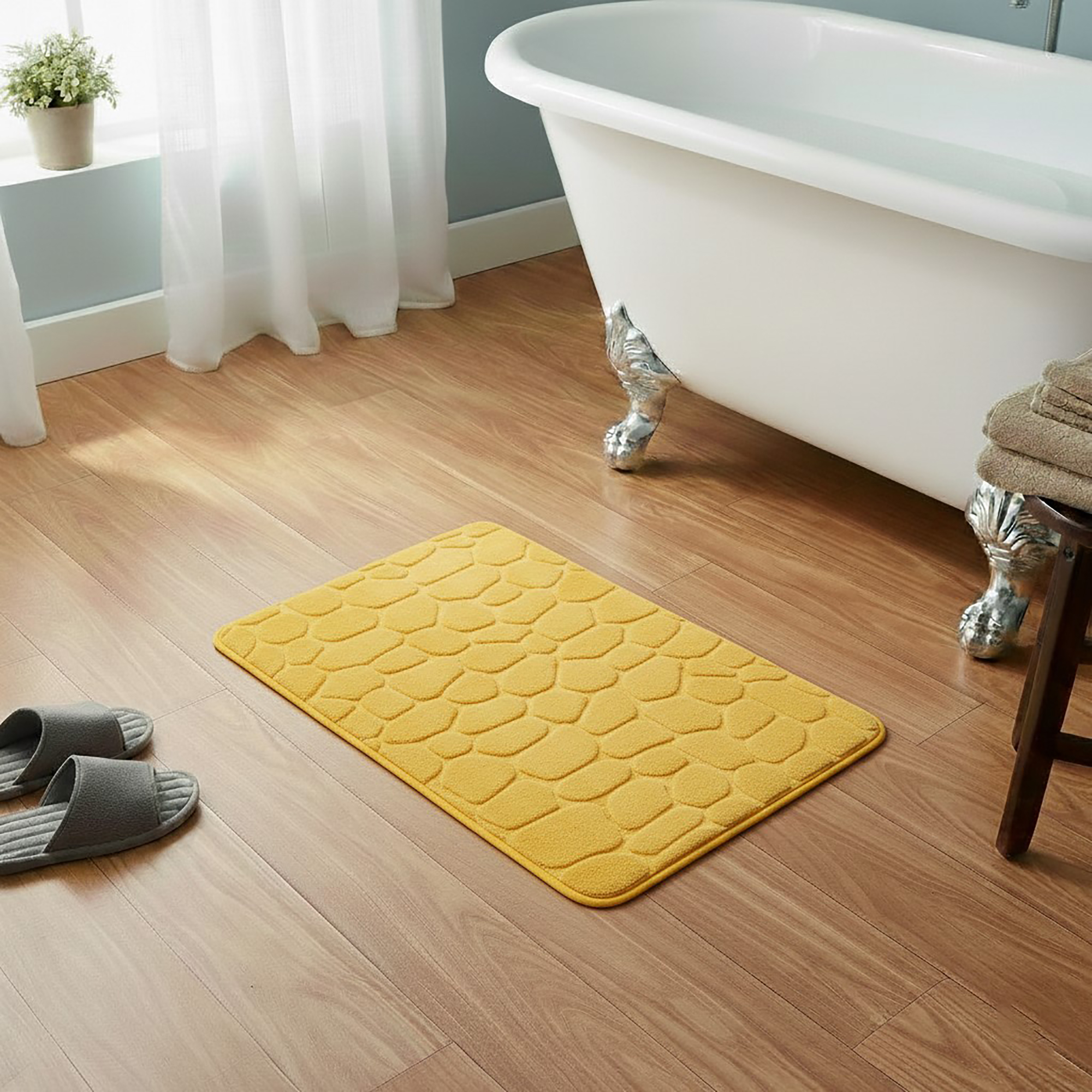 Mustard yellow memory foam bath mat with embossed pebble stone texture on wooden floor beside clawfoot tub