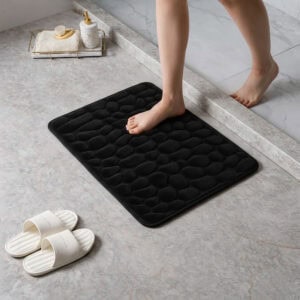 Person standing barefoot on a black memory foam mat with pebble texture placed on a bathroom tile floor